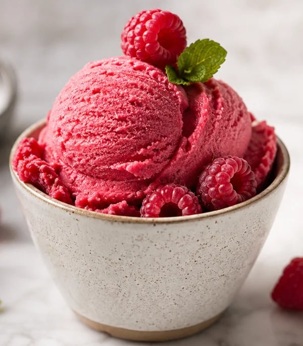 Pink raspberry ice cream scoops in a speckled ceramic bowl, topped with fresh raspberries and mint leaf