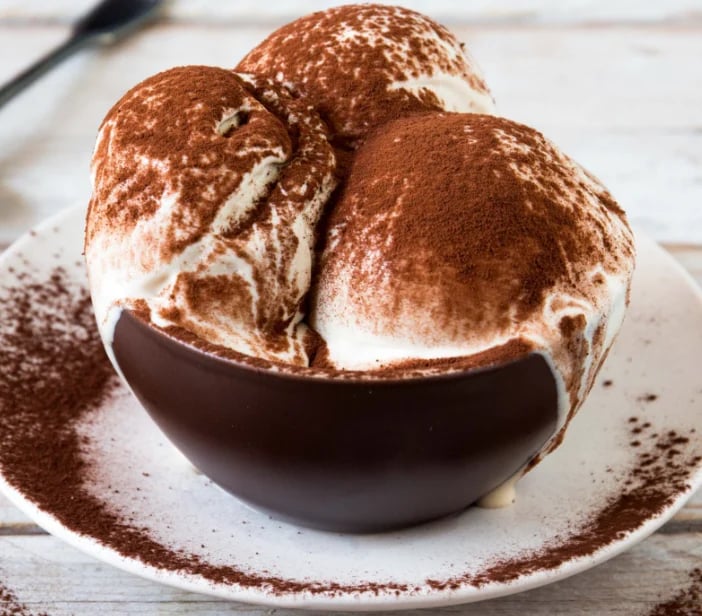 Chocolate tiramisu dessert dusted with cocoa powder on a white plate with a wooden surface background